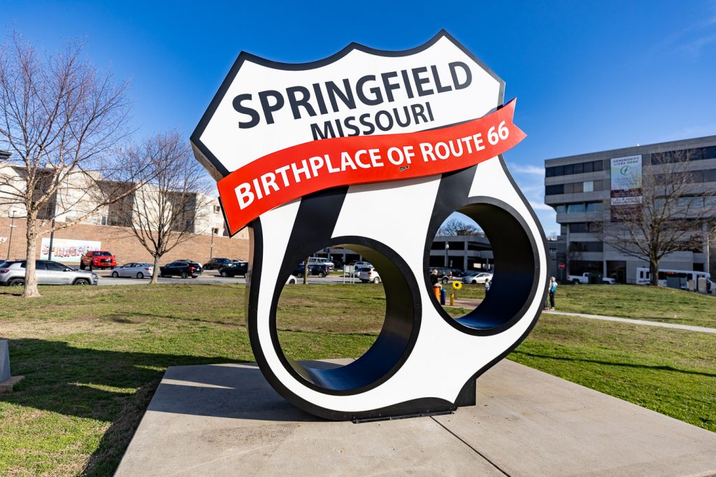 Large Black and White Route 66 shield with cutouts in the circles of the sixes for photos. Across the top it says "SPRINGFIELD, MISSOURI" with a red banner that says "BIRTHPLACE OF ROUTE 66." Birthplace of Route 66 Monument in Springfield, Missouri